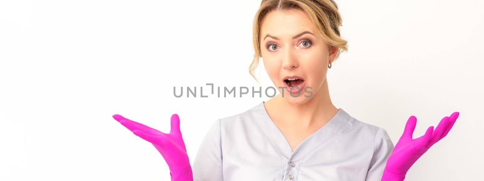 Body language. Young surprised Caucasian woman doctor wearing gloves gesturing with her hands, spread his arms having shocked expression opening her mouth against the white background