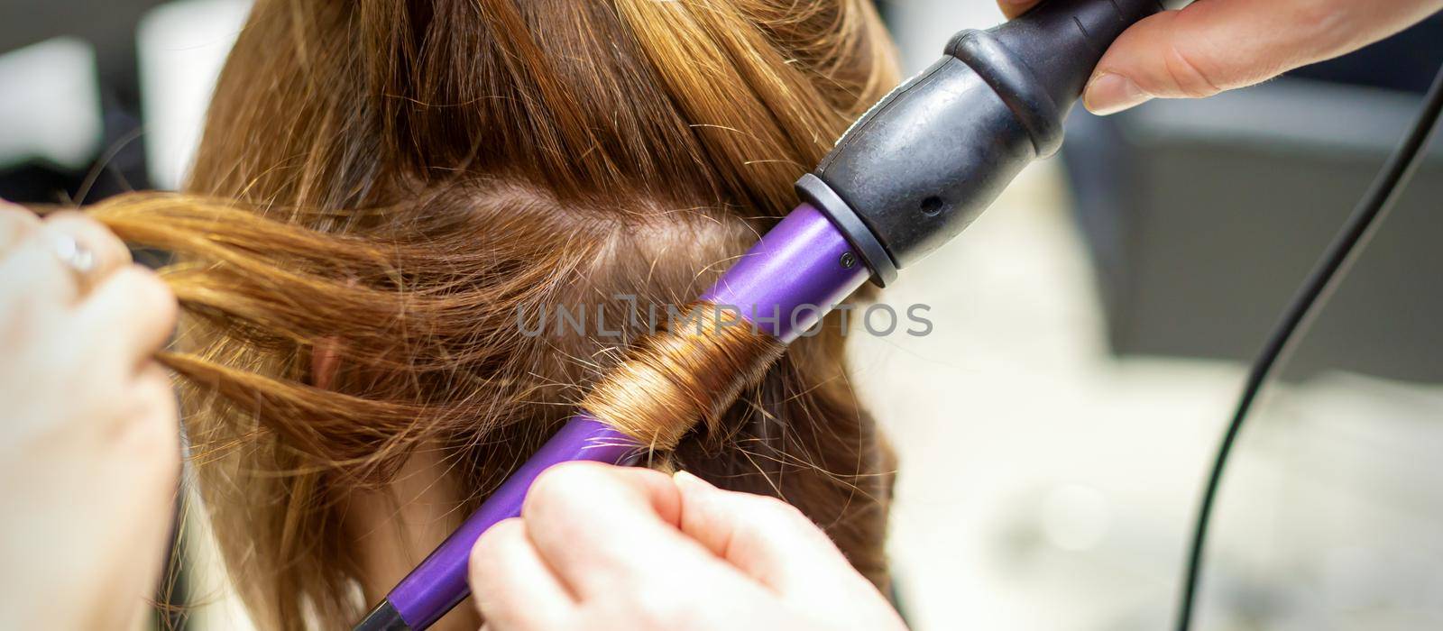 The hairstylist makes curls hairstyle of long brown hair with the curling iron in hairdresser salon, close up. by okskukuruza