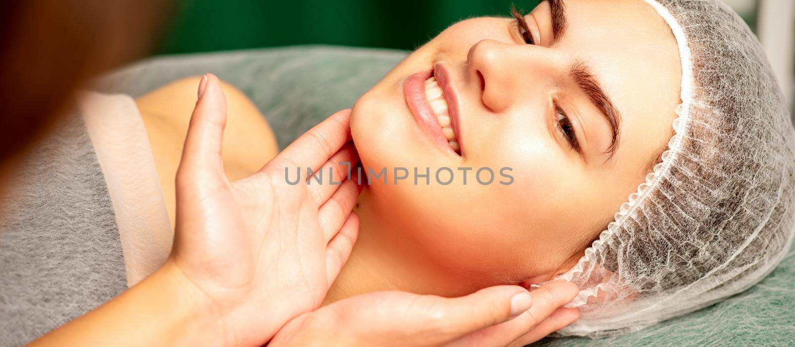 Plastic surgery and beauty concept. Professional cosmetician examining and touching the face of the beautiful young caucasian smiling woman in a clinic of esthetic cosmetology. by okskukuruza