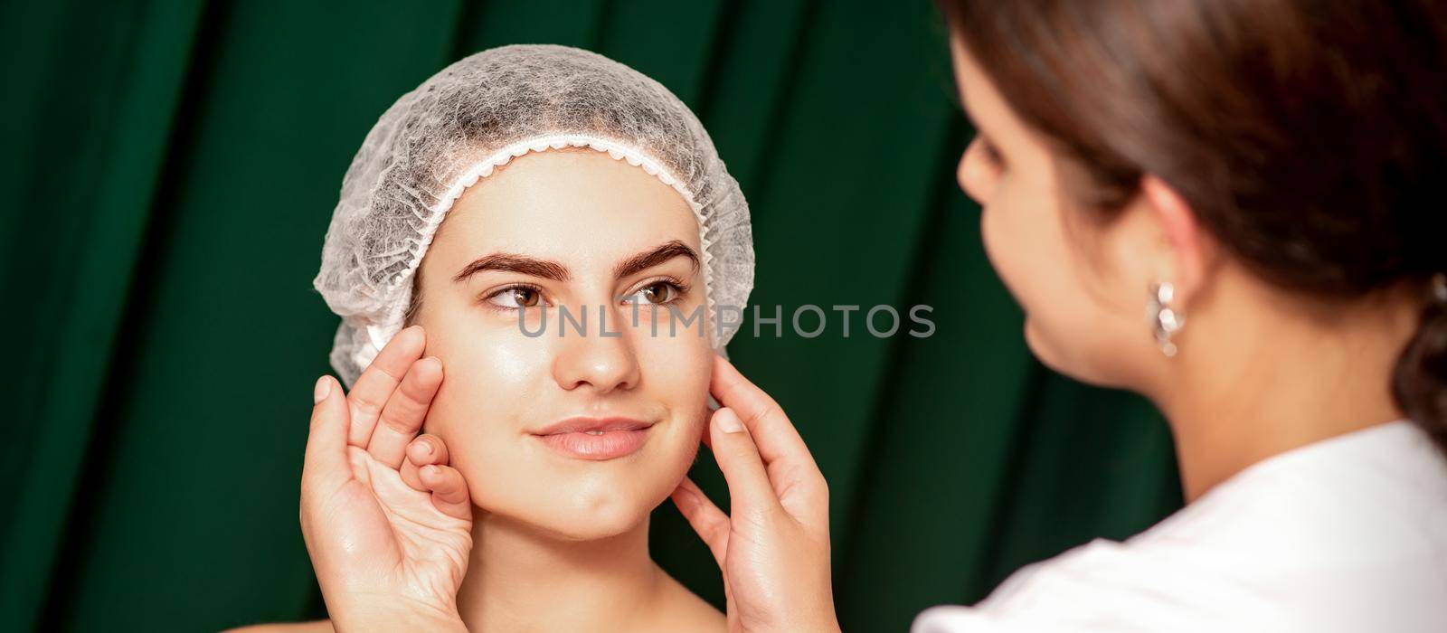 Plastic surgery and beauty concept. Professional cosmetician examining and touching the face of the beautiful young caucasian smiling woman in a clinic of esthetic cosmetology