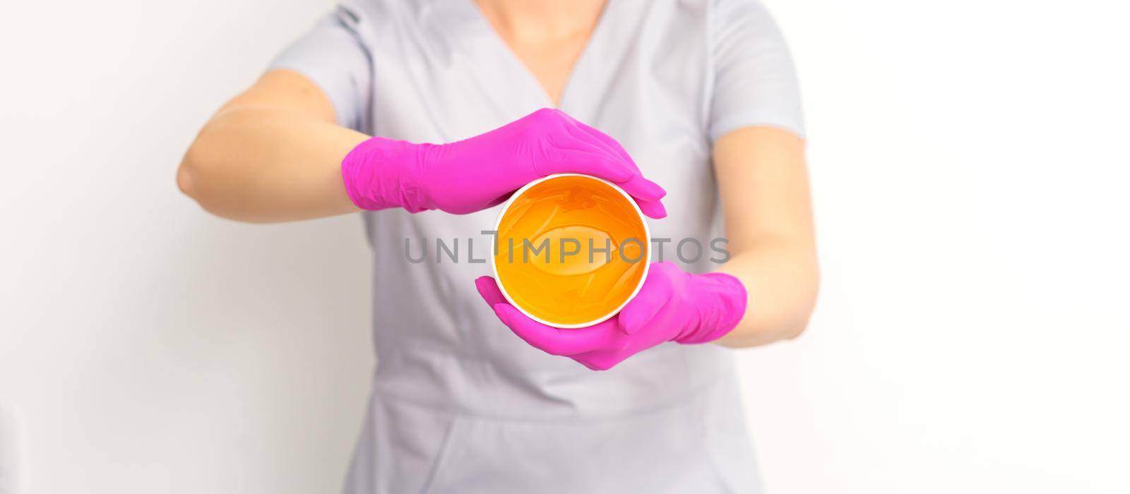 Portrait of a female caucasian beautician holding a jar of sugar paste for sugaring wearing pink gloves on white background. by okskukuruza