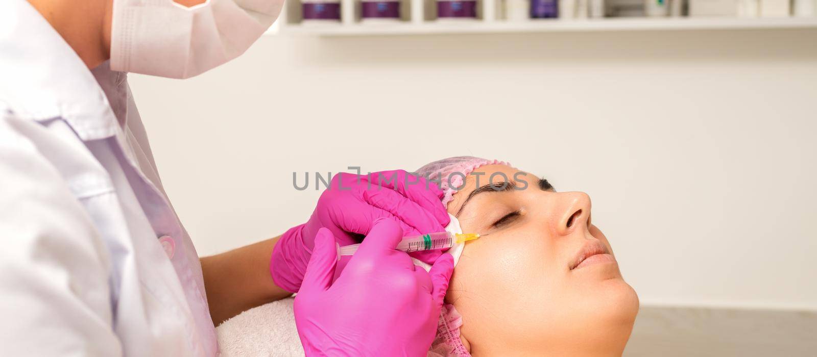 The young white woman is getting rejuvenating facial injections with hyaluronic acid on the eye in a beauty clinic