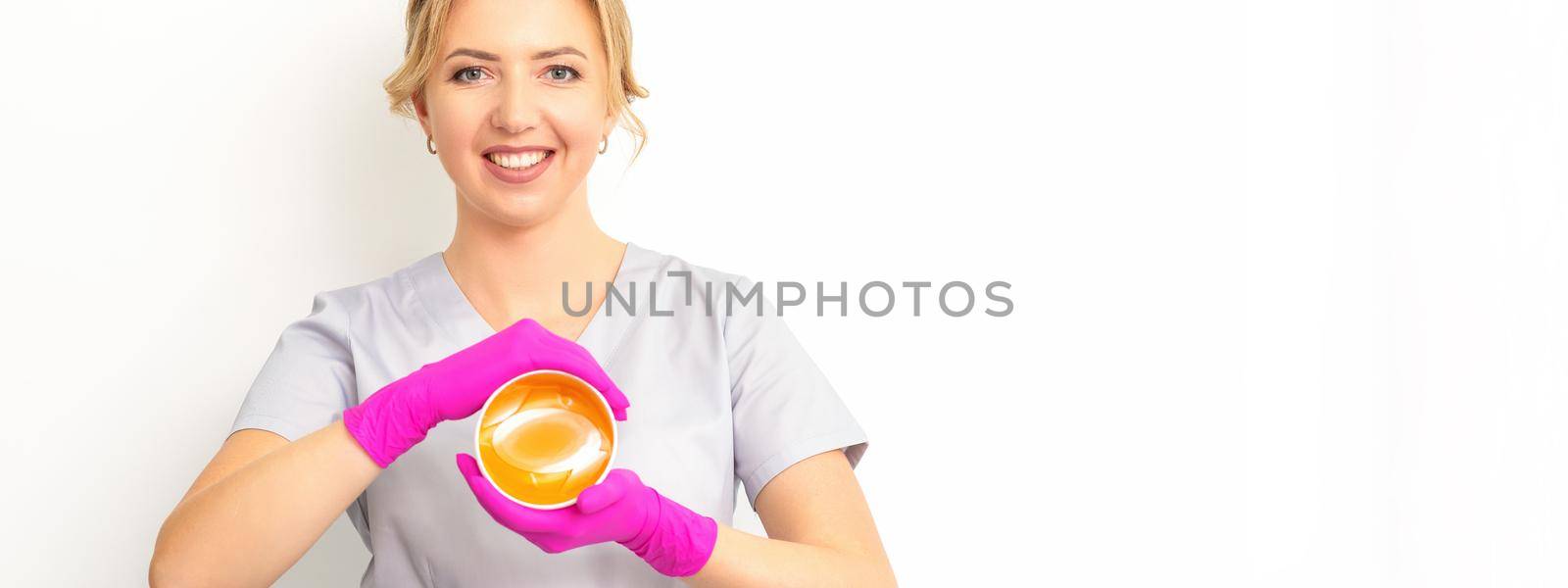 Portrait of a female caucasian beautician holding a jar of sugar paste for sugaring wearing pink gloves on white background. by okskukuruza