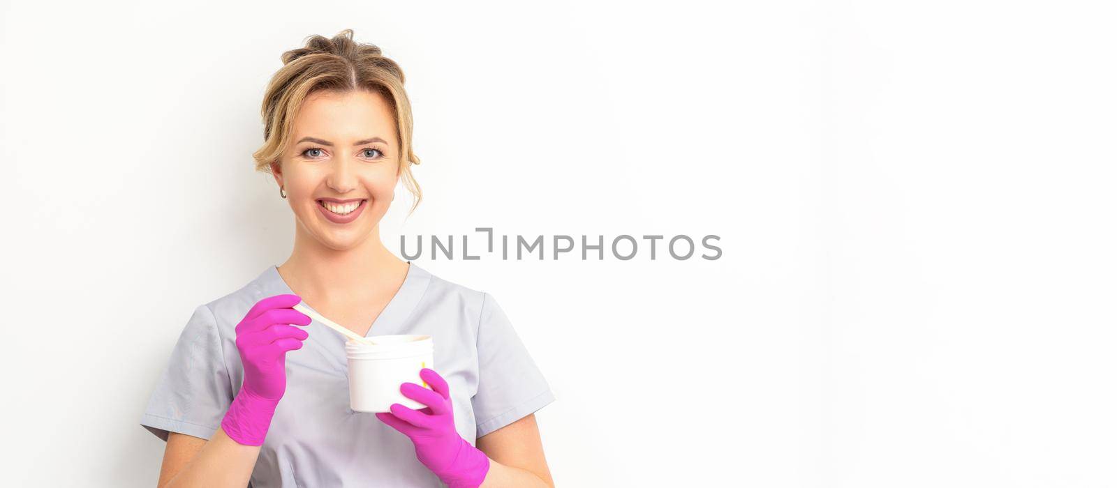 Portrait of a female caucasian beautician holding a jar of sugar paste for sugaring wearing pink gloves on white background