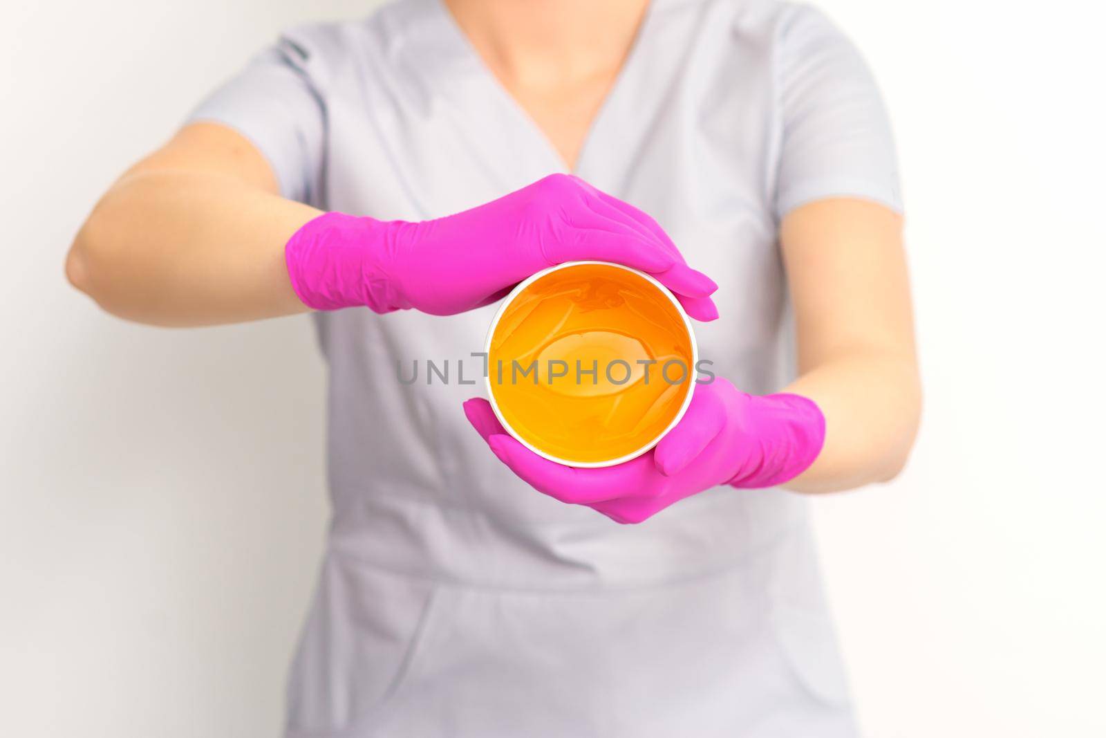 Portrait of a female caucasian beautician holding a jar of sugar paste for sugaring wearing pink gloves on white background