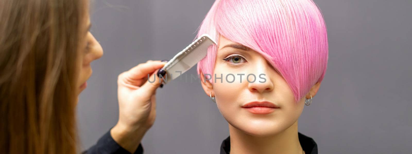 The female hairdresser is combing the dyed pink short hair of the young woman against the dark wall