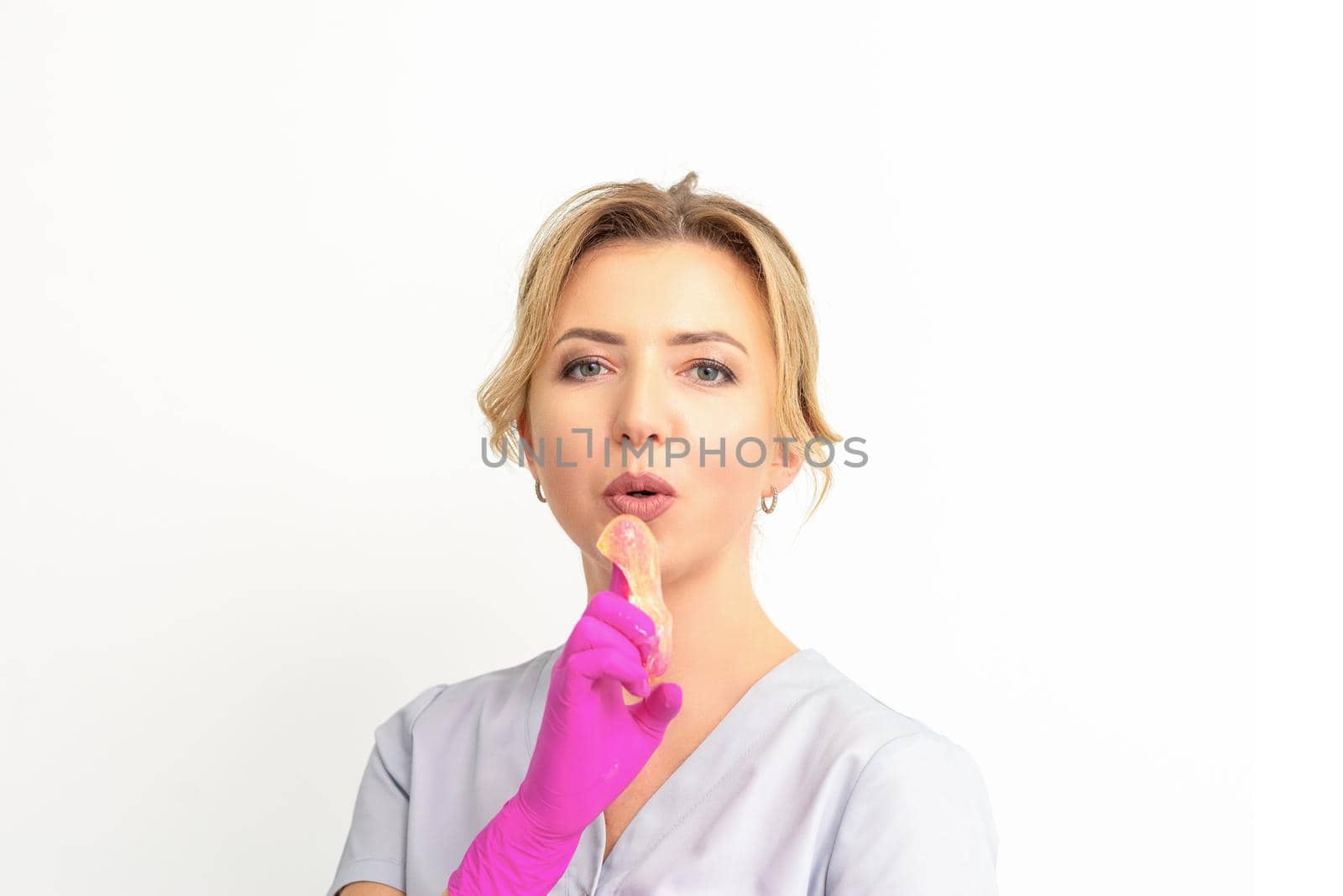 Young caucasian female beautician holding wax paste on forefinger blowing on him wearing pink gloves against a white background. Epilation concept