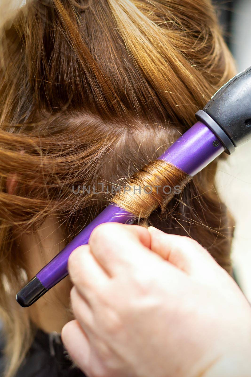The hairstylist makes curls hairstyle of long brown hair with the curling iron in hairdresser salon, close up