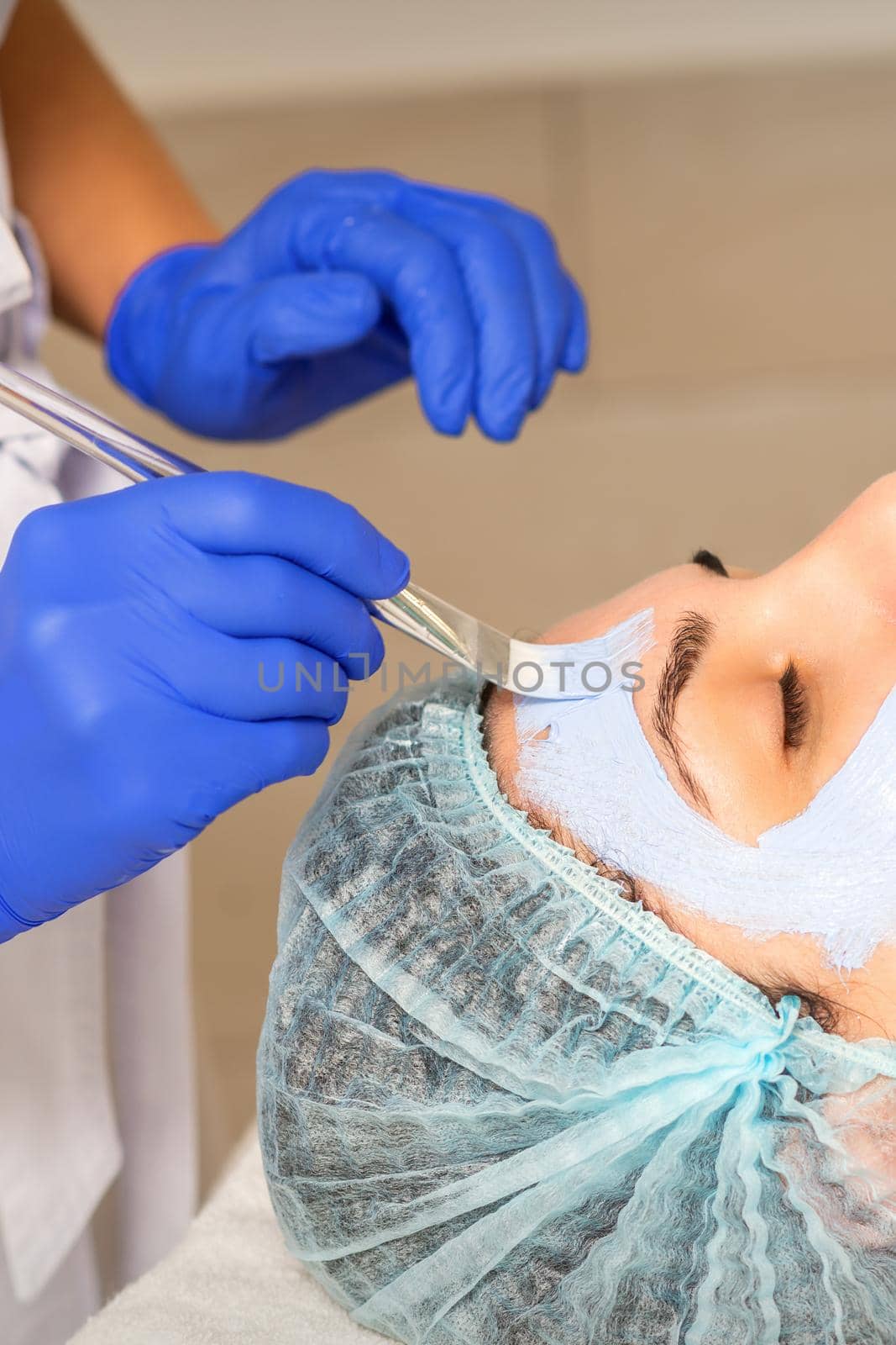 The beautician with brush applies a photochemical and glycolic peeling face mask to the female patient face in the beauty clinic