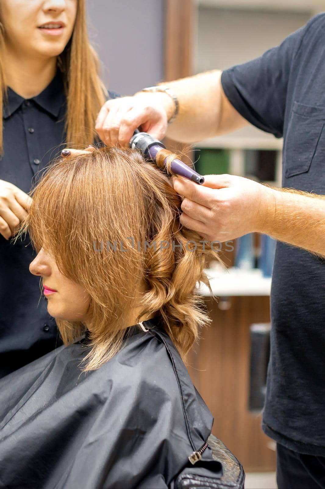 Two hairstylists make curls hairstyle of long brown hair with the curling iron in hairdresser salon. by okskukuruza