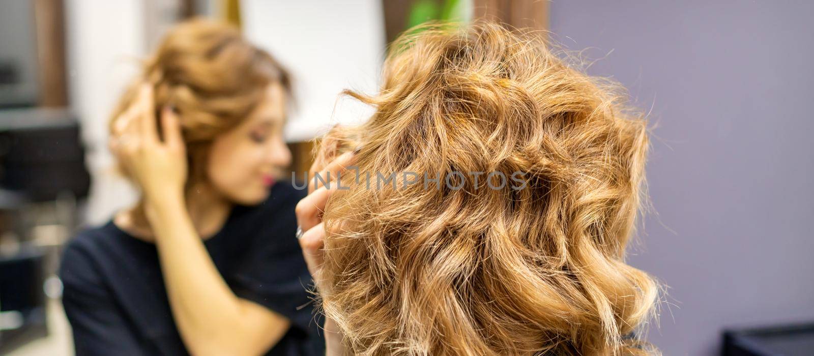 Young woman checking her new curly brown hairstyle in front of the mirror at the hairdresser salon.