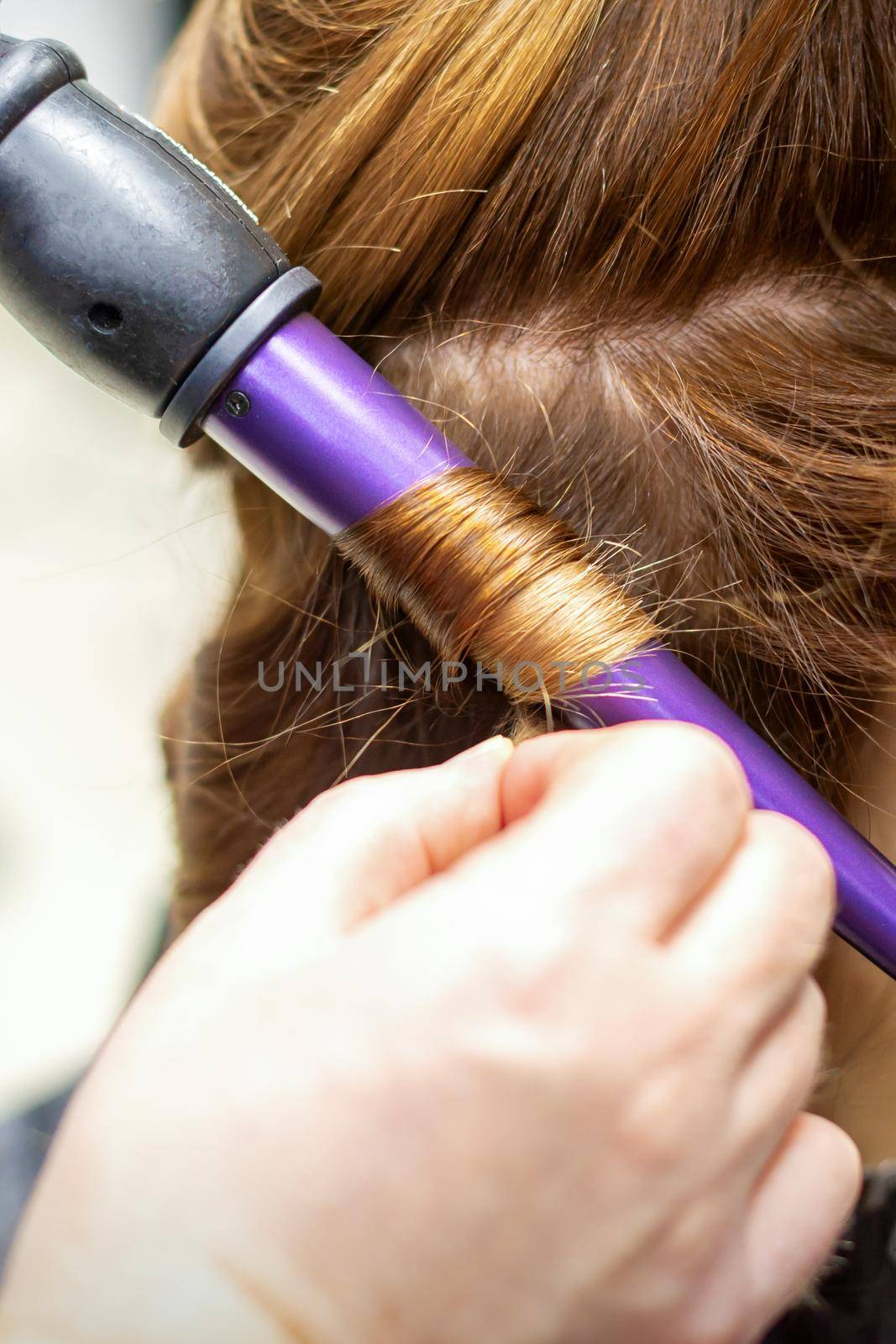 The hairstylist makes curls hairstyle of long brown hair with the curling iron in hairdresser salon, close up