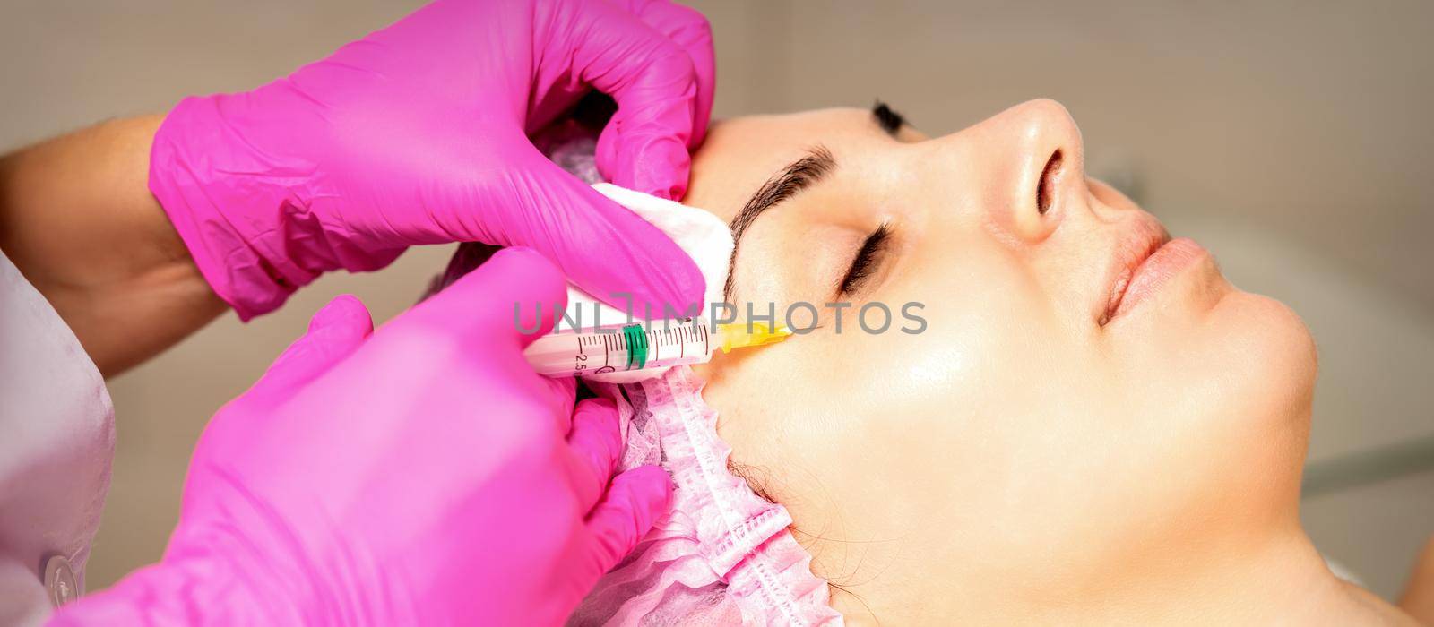 The young white woman is getting rejuvenating facial injections with hyaluronic acid on the eye in a beauty clinic