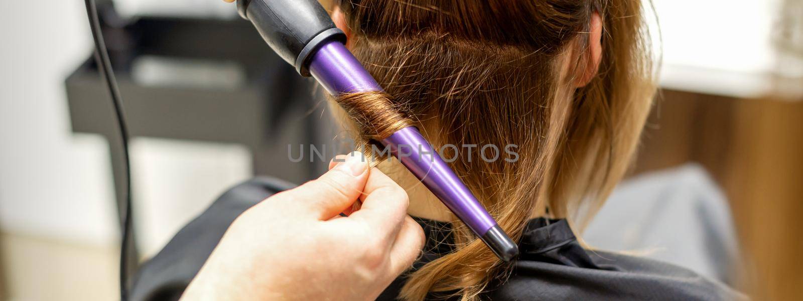 The hairstylist makes curls hairstyle of long brown hair with the curling iron in hairdresser salon, close up. by okskukuruza
