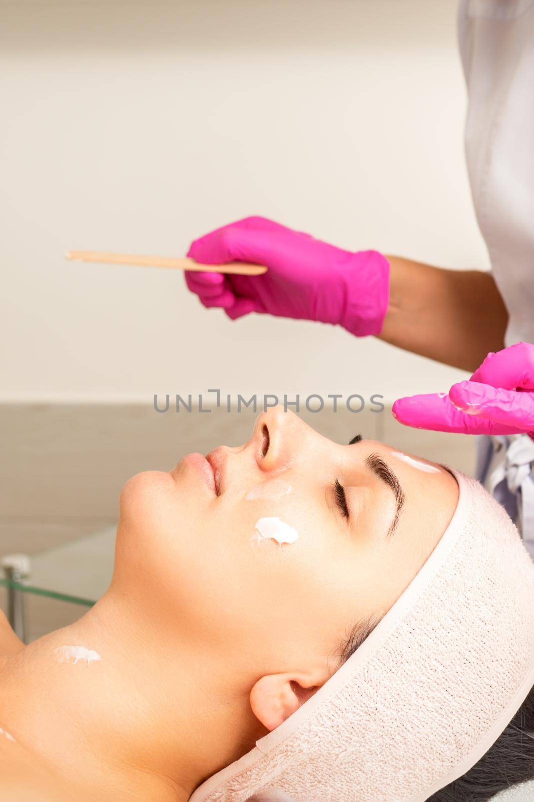 Cosmetologist with gloved hands applies a moisturizing mask with peeling cream on the female face. Facial cosmetology treatment. Procedures for facial care