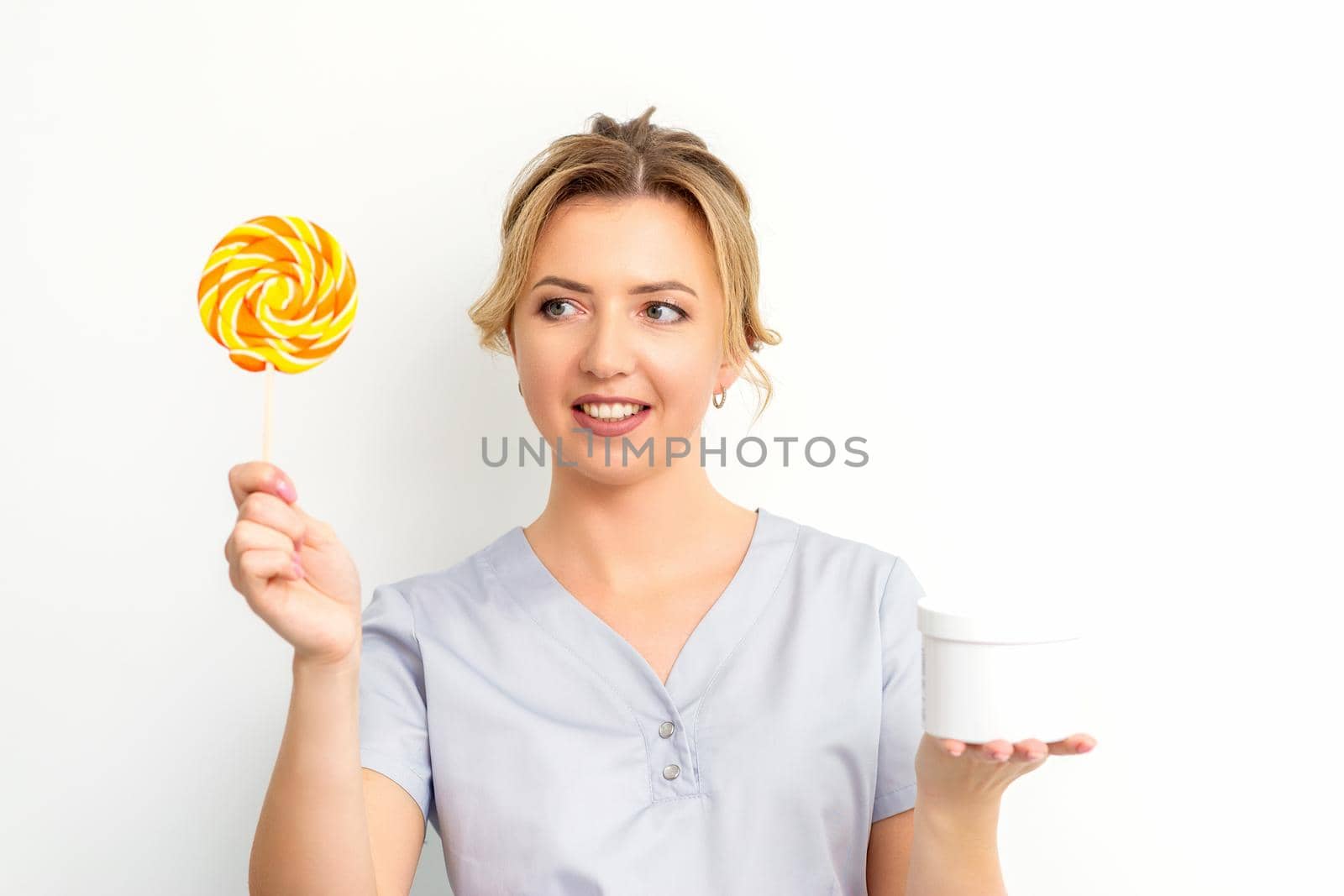 Waxing, depilation concept. The beautician is wearing a medical coat holding a sugar lollipop with a jar of sugar paste on white background. by okskukuruza