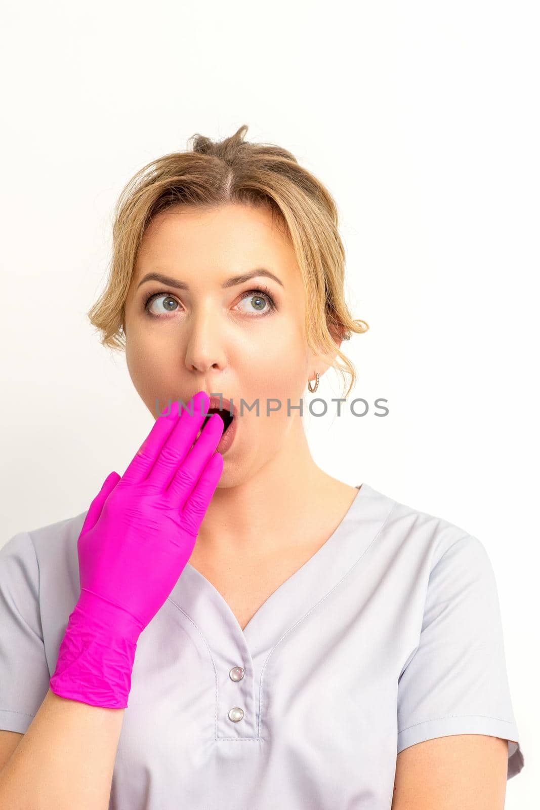 Portrait of a young female caucasian doctor or nurse is shocked covering her mouth with her pink gloved hands against a white background. by okskukuruza