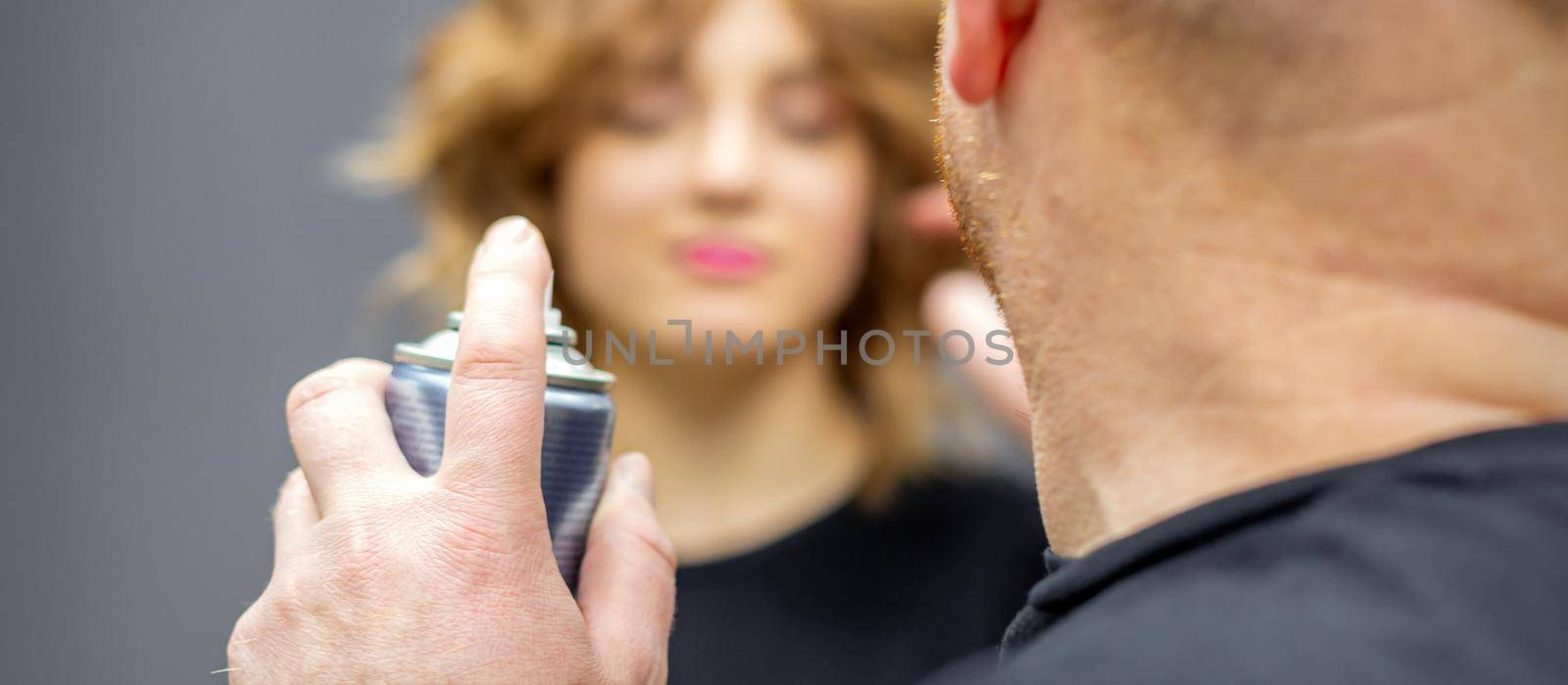 The hairdresser is using hair spray to fix the hairstyle of the young caucasian woman in the hairdresser salon, close up