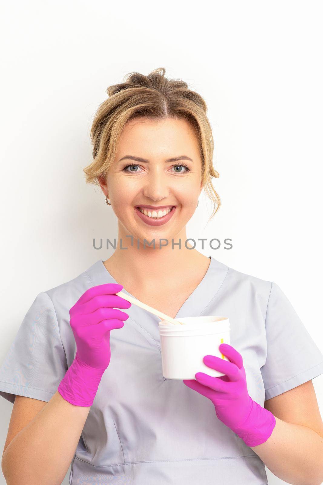 Portrait of a female caucasian beautician holding a jar of sugar paste for sugaring wearing pink gloves on white background