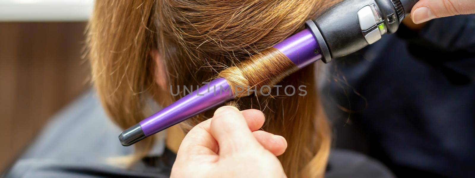 The hairstylist makes curls hairstyle of long brown hair with the curling iron in hairdresser salon, close up