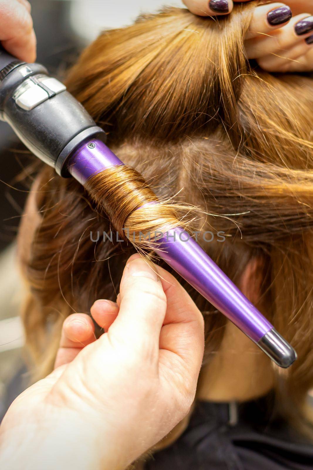 The hairstylist makes curls hairstyle of long brown hair with the curling iron in hairdresser salon, close up