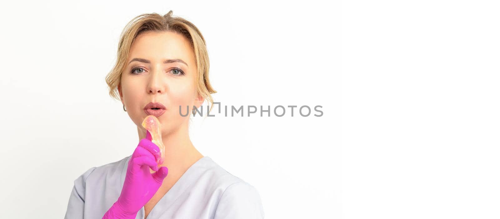 Young caucasian female beautician holding wax paste on forefinger blowing on him wearing pink gloves against a white background. Epilation concept