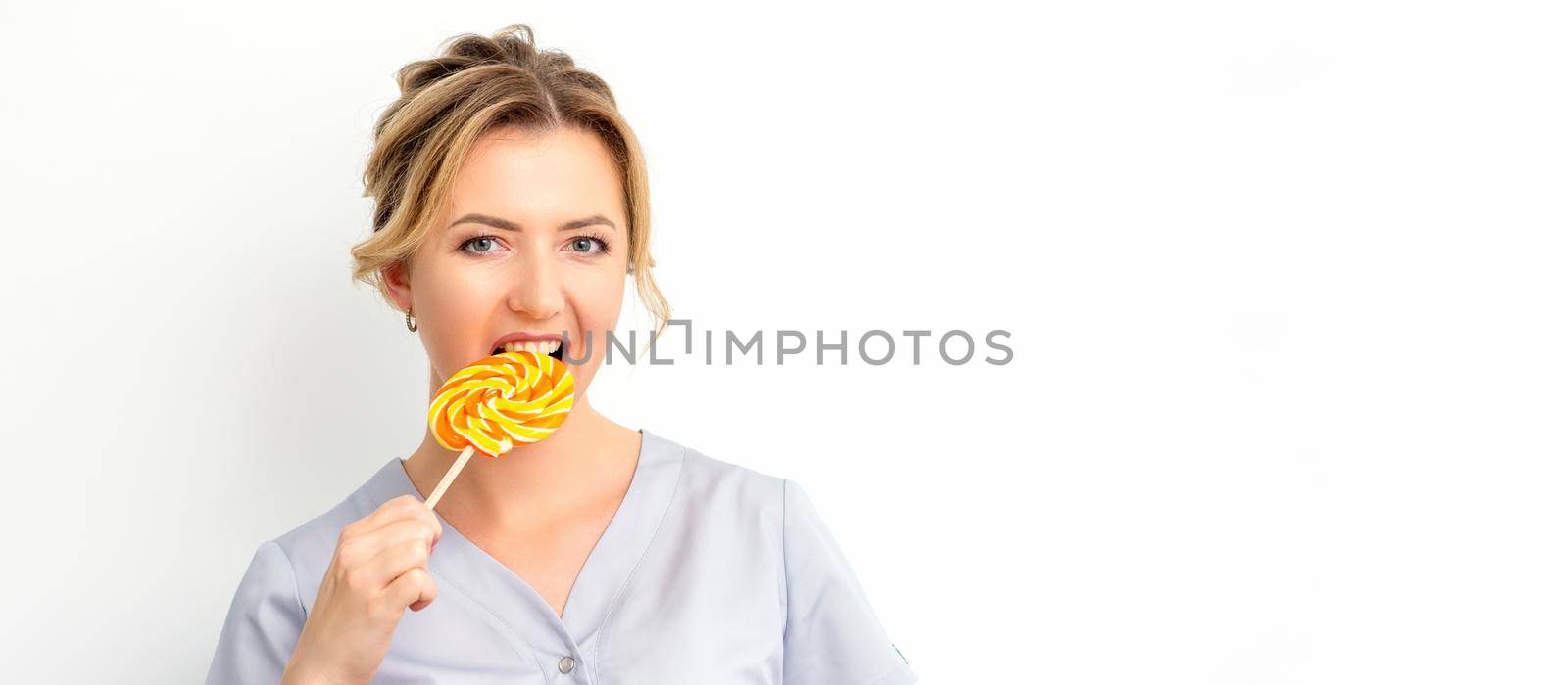 Portrait of a beautiful young caucasian beautician wearing a medical shirt bites a lollipop on a white background