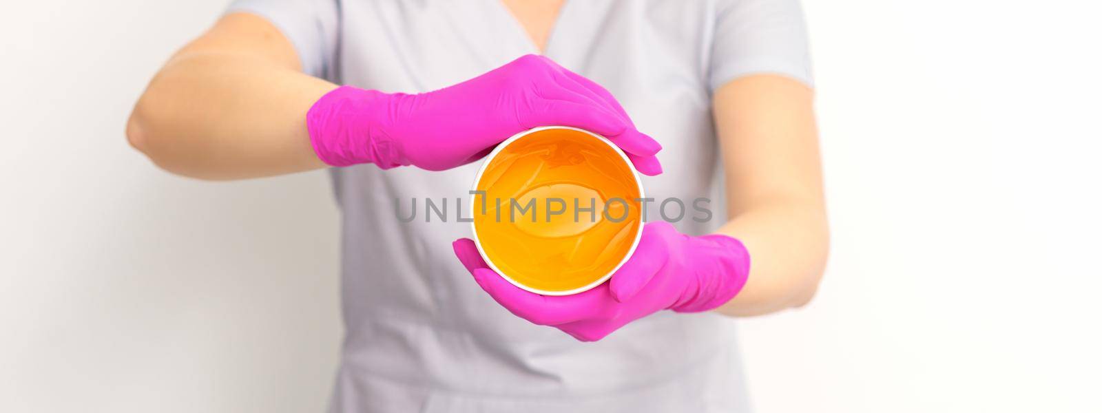 Portrait of a female caucasian beautician holding a jar of sugar paste for sugaring wearing pink gloves on white background