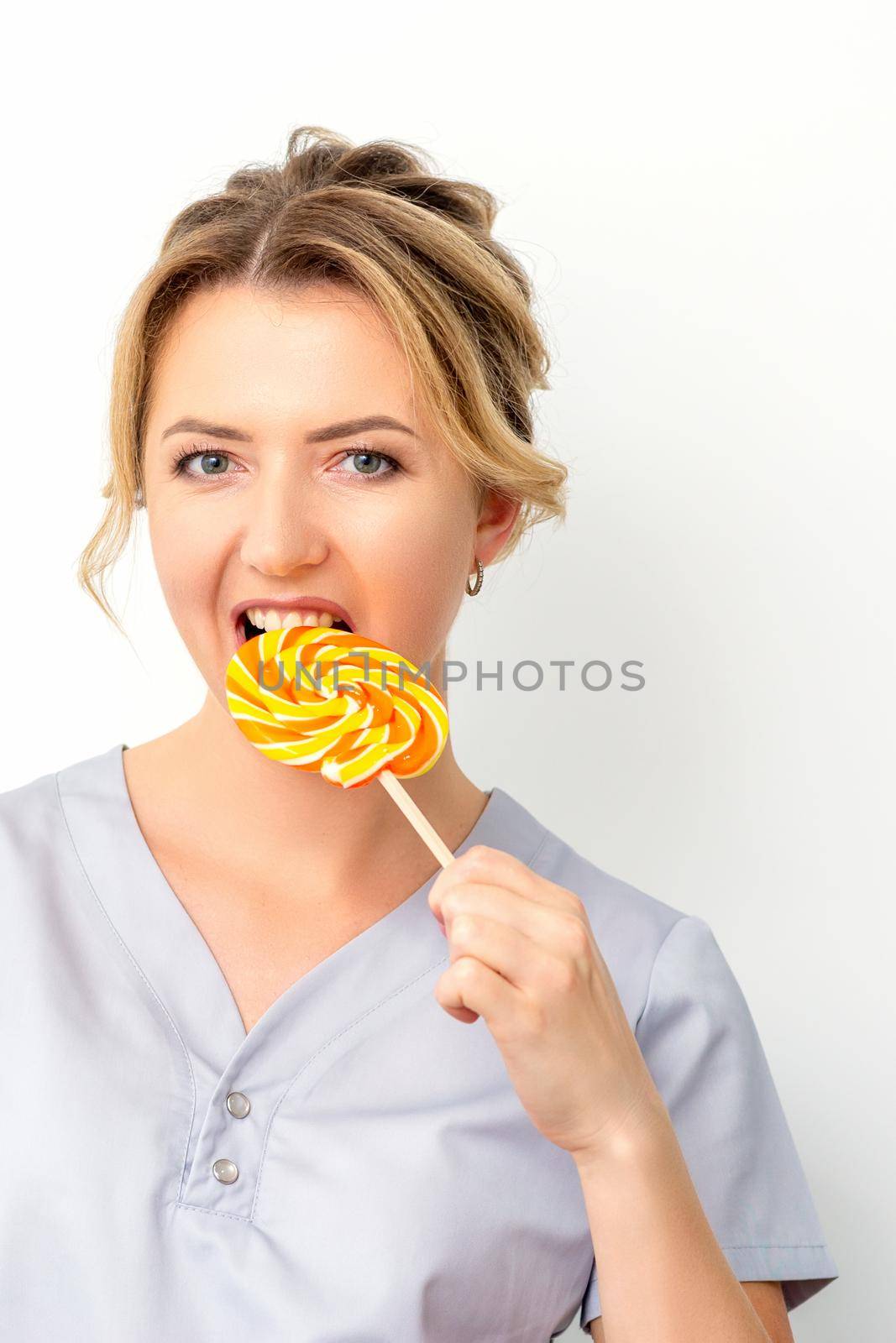 Portrait of a beautiful young caucasian beautician wearing a medical shirt bites a lollipop on a white background. by okskukuruza