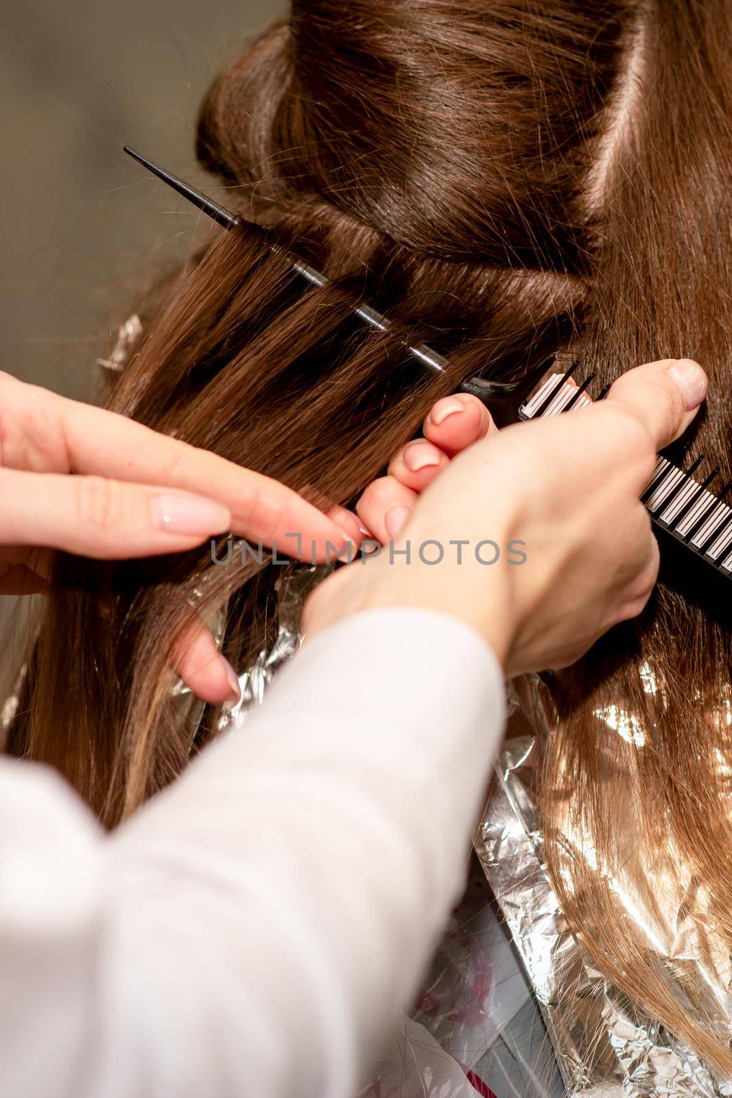 Hairdresser's hands prepare brown hair for dyeing with a comb and foil in a beauty salon, close up. by okskukuruza