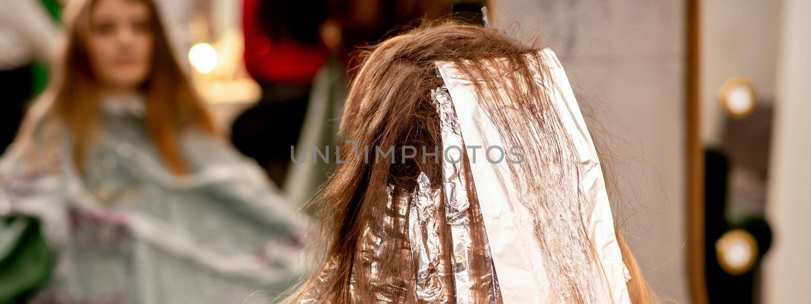 Beautiful young caucasian woman with foil in her hair while dyeing her hair sitting in a beauty salon. by okskukuruza