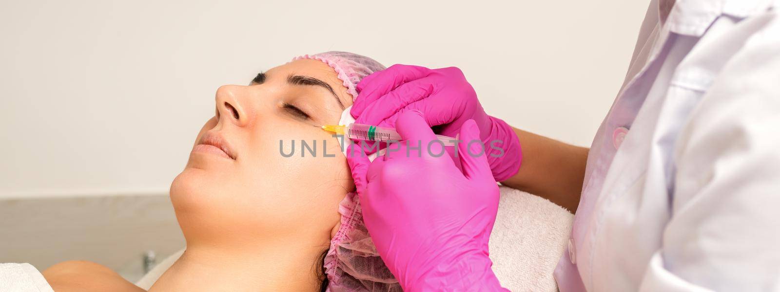 The young white woman is getting rejuvenating facial injections with hyaluronic acid on the eye in a beauty clinic