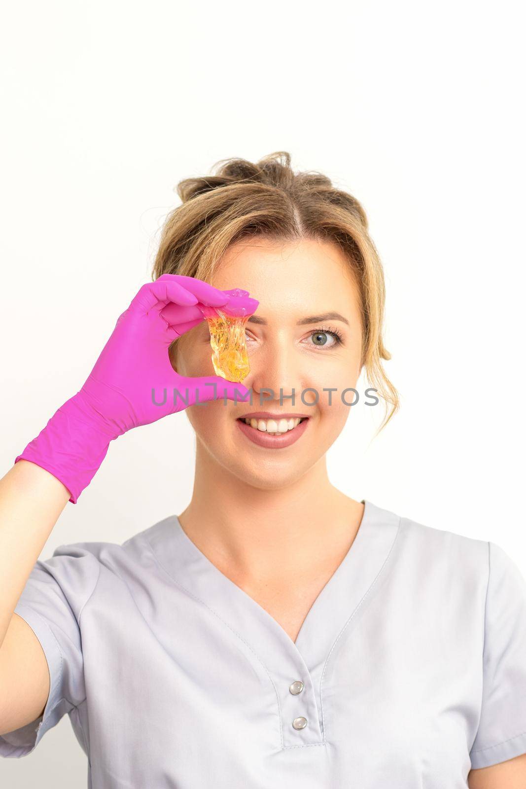 The master of sugar hair removal holds liquid yellow sugar paste, covering her eye with wax for depilation on a white background