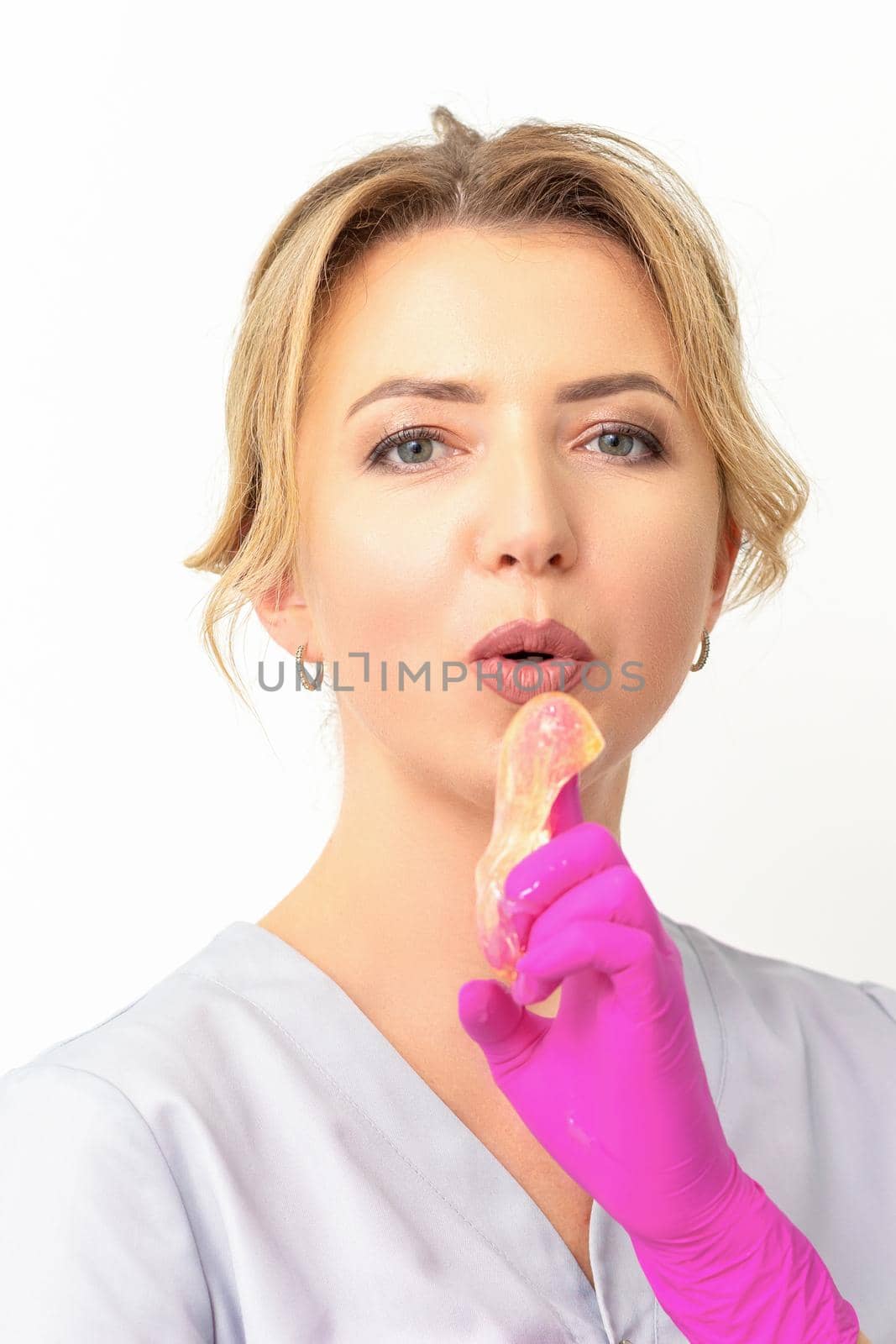 Young caucasian female beautician holding wax paste on forefinger blowing on him wearing pink gloves against a white background. Epilation concept. by okskukuruza