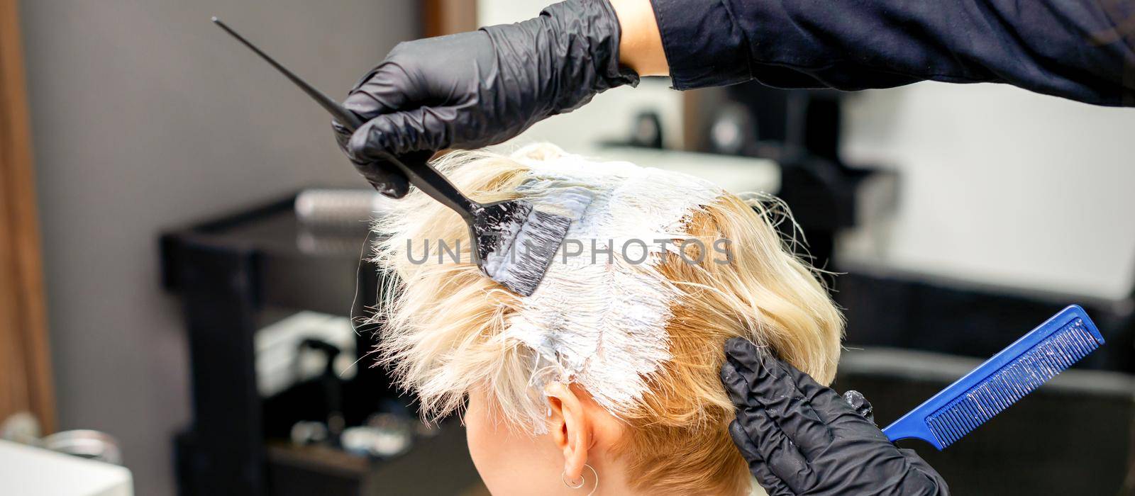 Coloring white hair with hair dye and brush by hands of hairstylist for the young caucasian blonde woman at a hair salon, close up