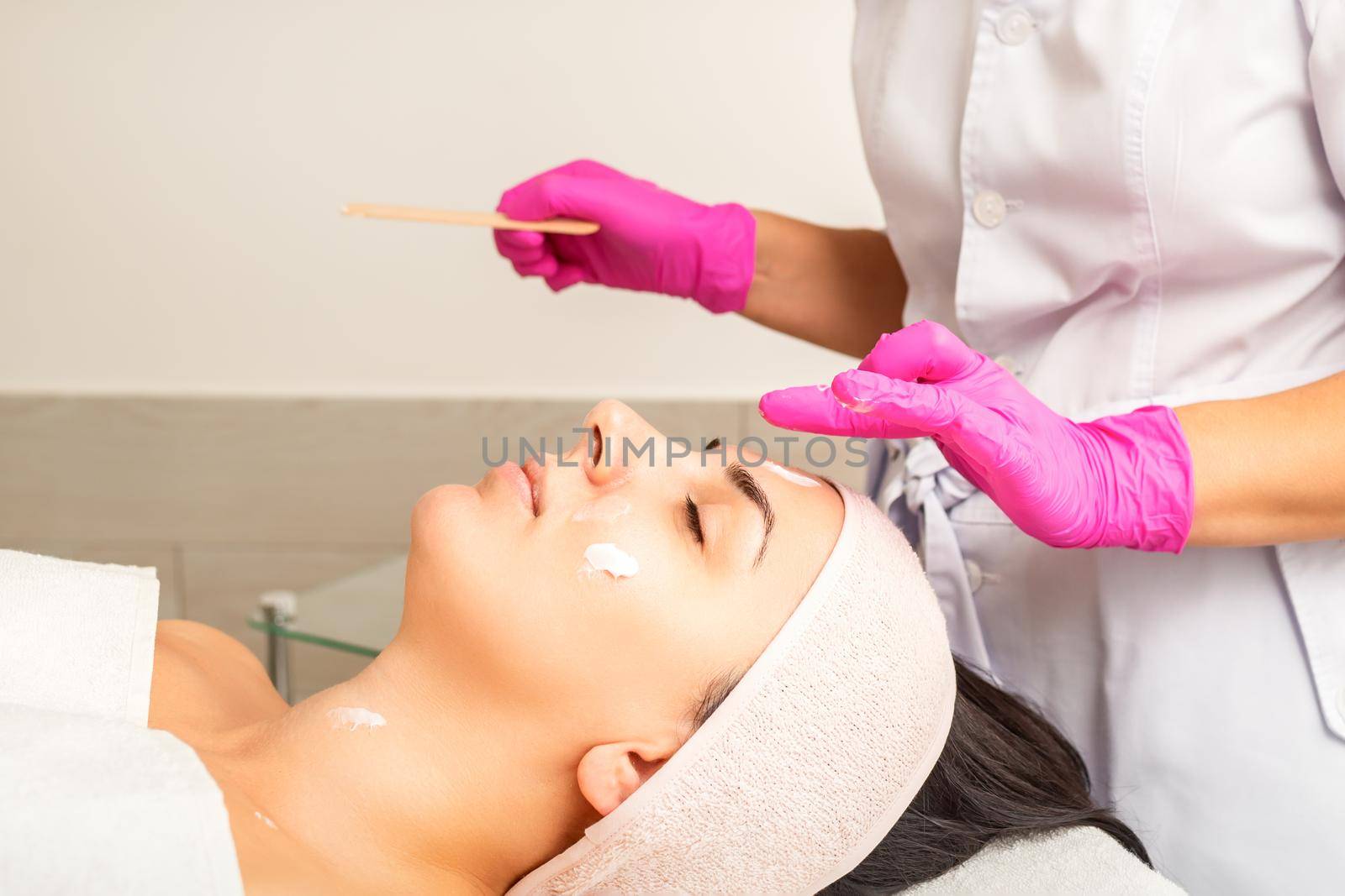 Cosmetologist with gloved hands applies a moisturizing mask with peeling cream on the female face. Facial cosmetology treatment. Procedures for facial care