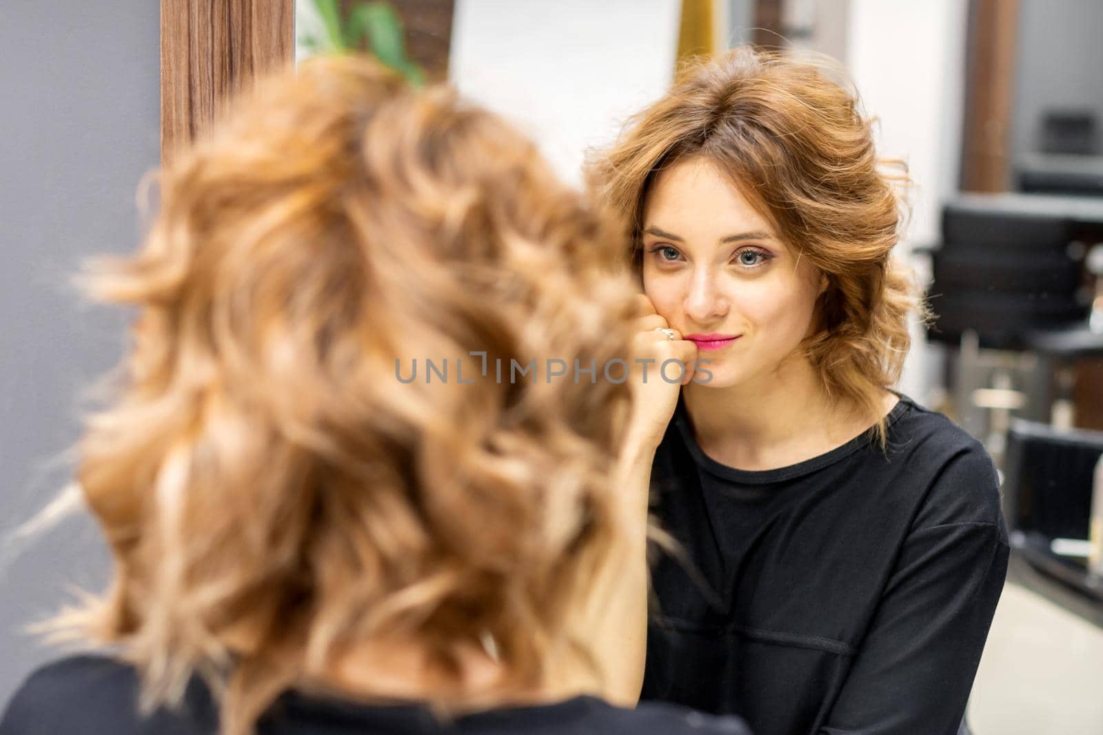 The beautiful young caucasian red-haired woman with a new short wavy hairstyle looking in the mirror checking hairstyle and makeup in a hairdresser salon. by okskukuruza