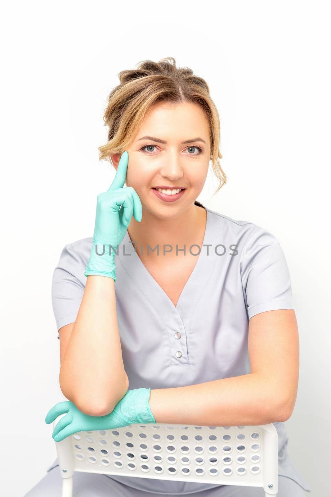 Close-up portrait of young smiling female caucasian healthcare worker sitting and staring at the camera wearing gloves on white background