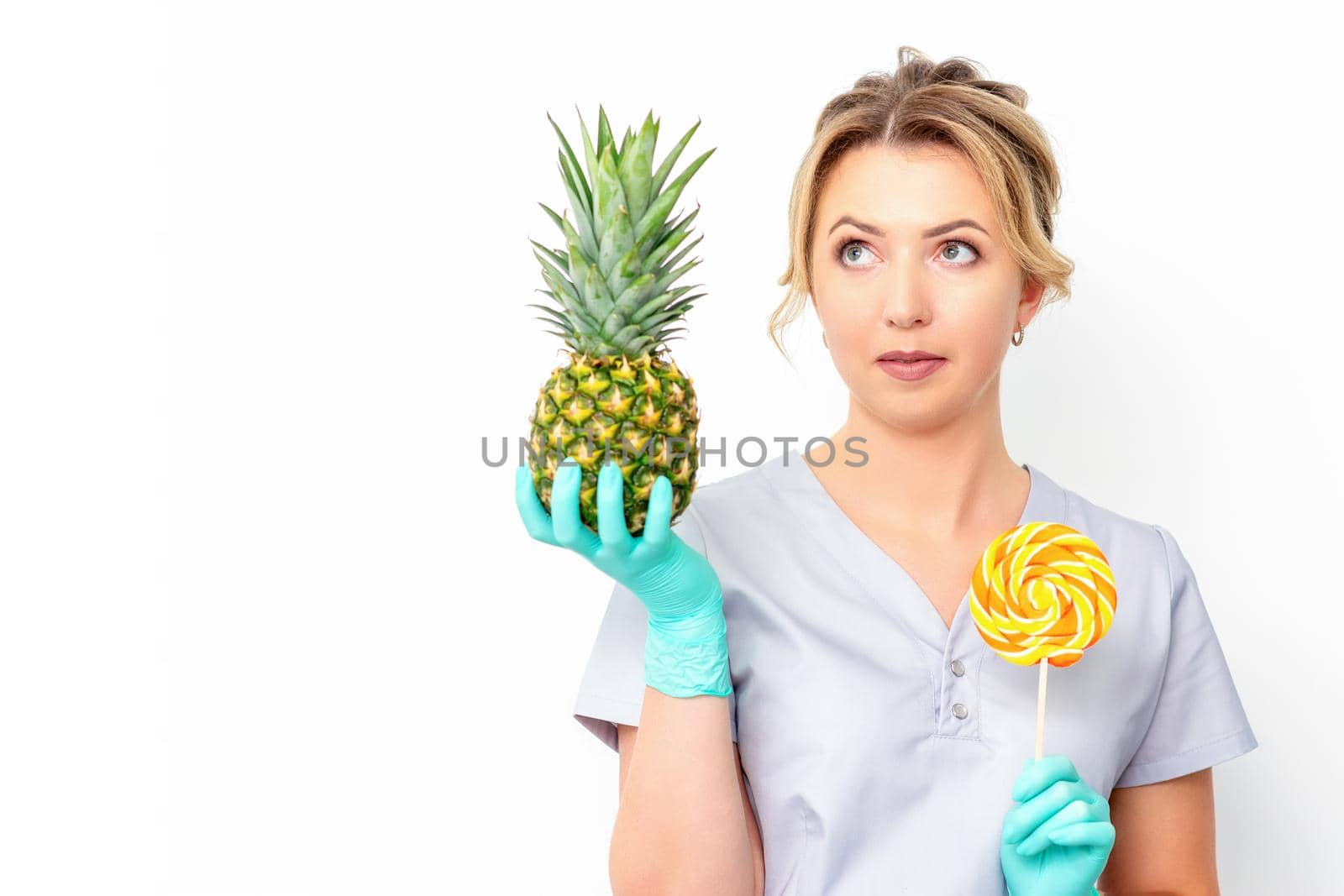 Young caucasian female doctor nutritionist holding fresh pineapple with lollipop over white background. by okskukuruza
