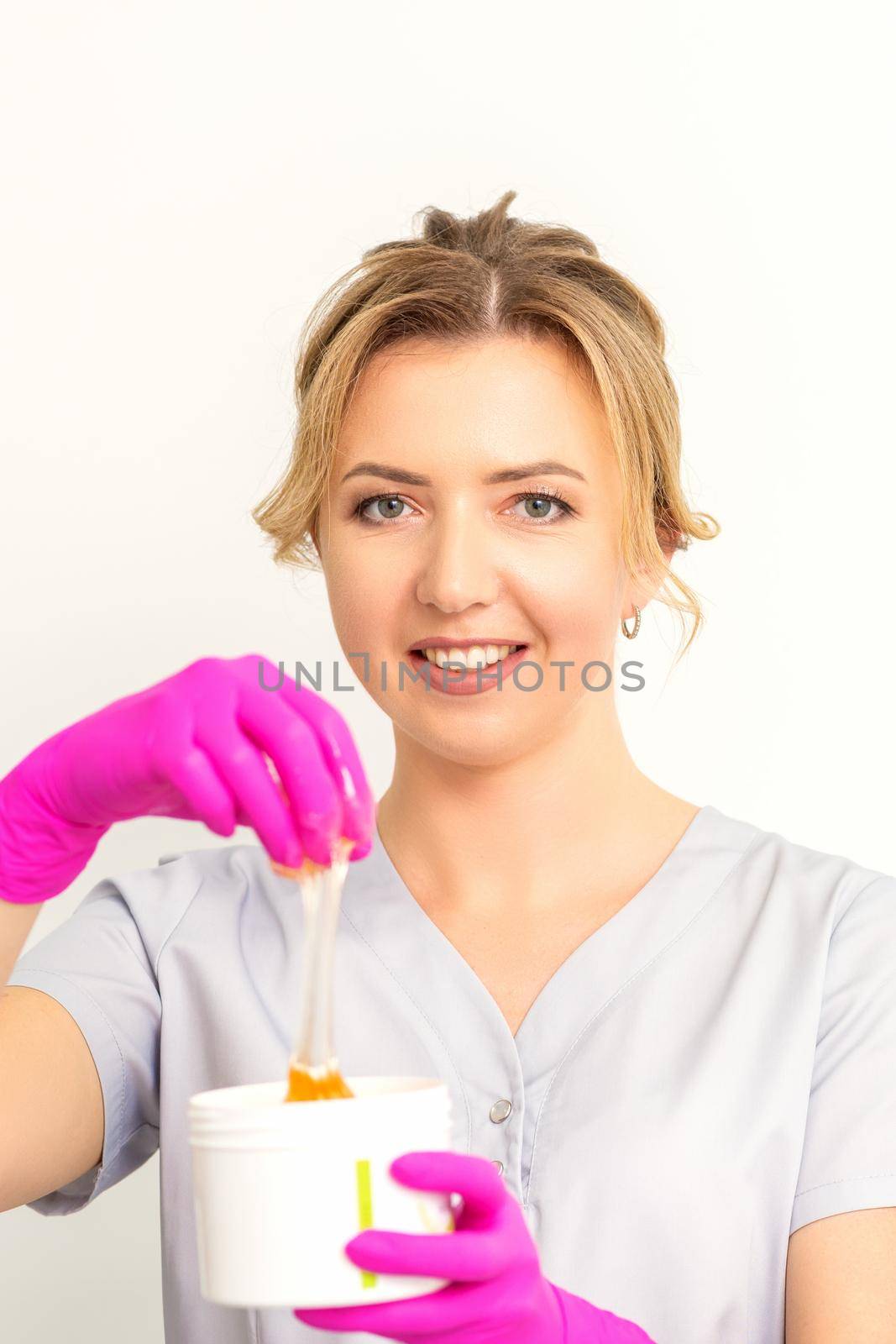 Portrait of a female caucasian beautician holding a jar of sugar paste for sugaring wearing pink gloves on white background. by okskukuruza