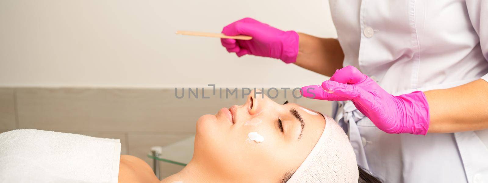 Cosmetologist with gloved hands applies a moisturizing mask with peeling cream on the female face. Facial cosmetology treatment. Procedures for facial care