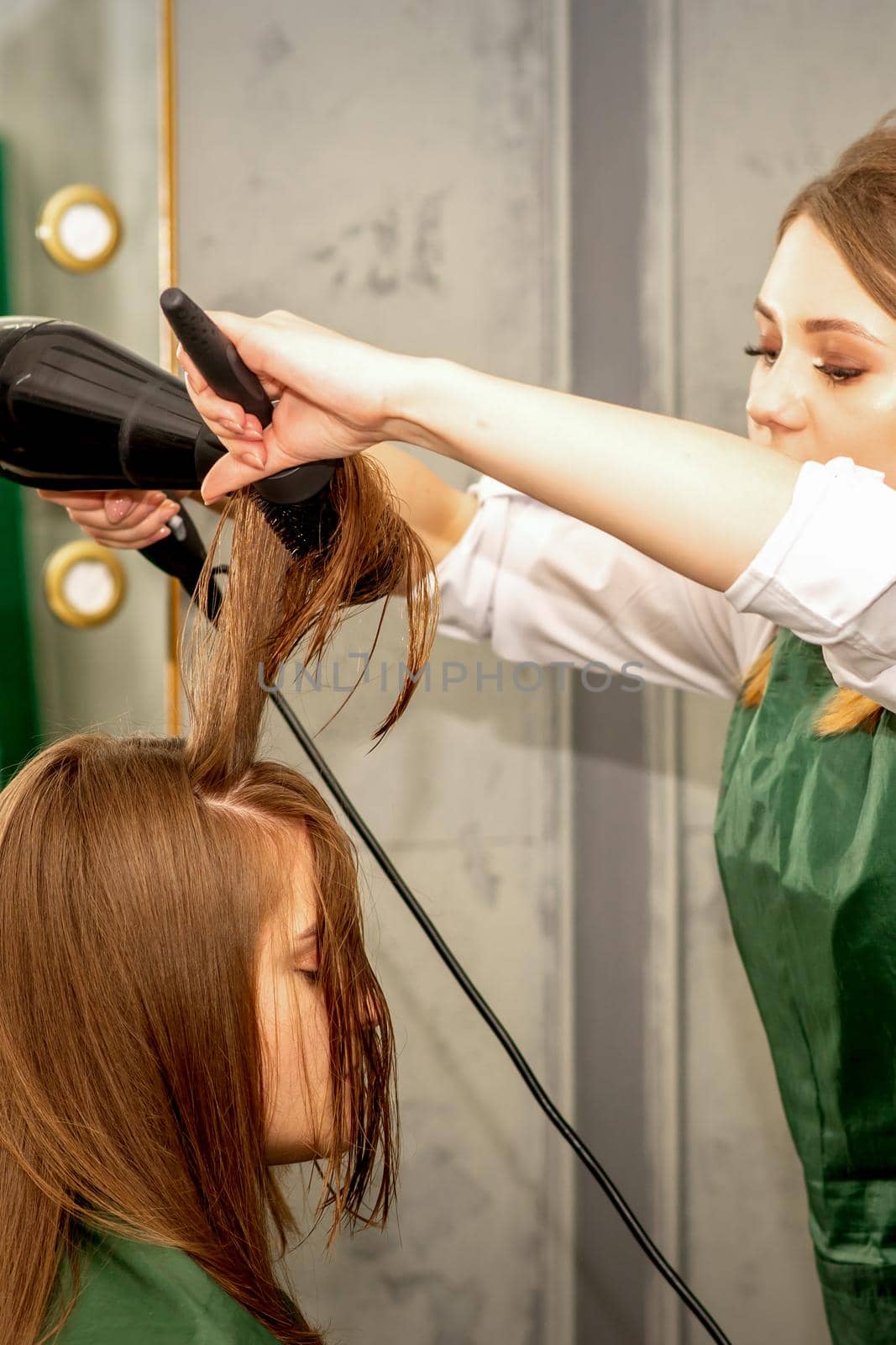 The hairdresser is drying long brown hair with a hairdryer and round brush in a beauty salon