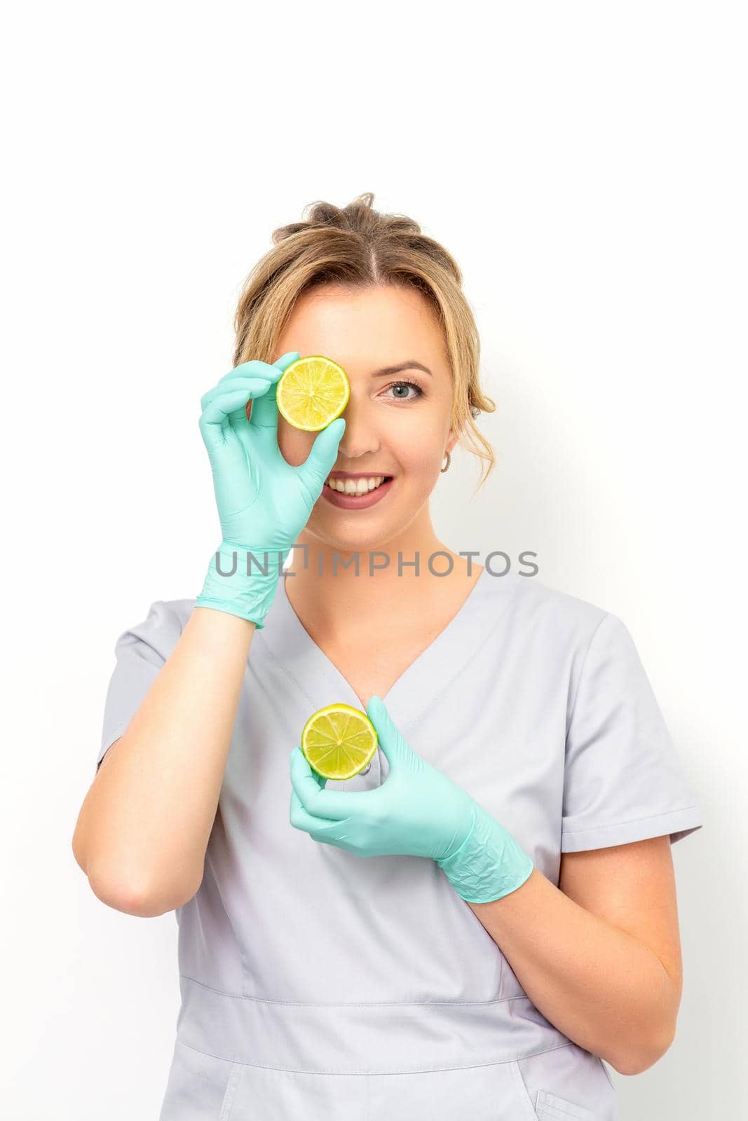 Portrait of young caucasian smiling female beautician covering eye with a lime slice wearing gloves over a white background