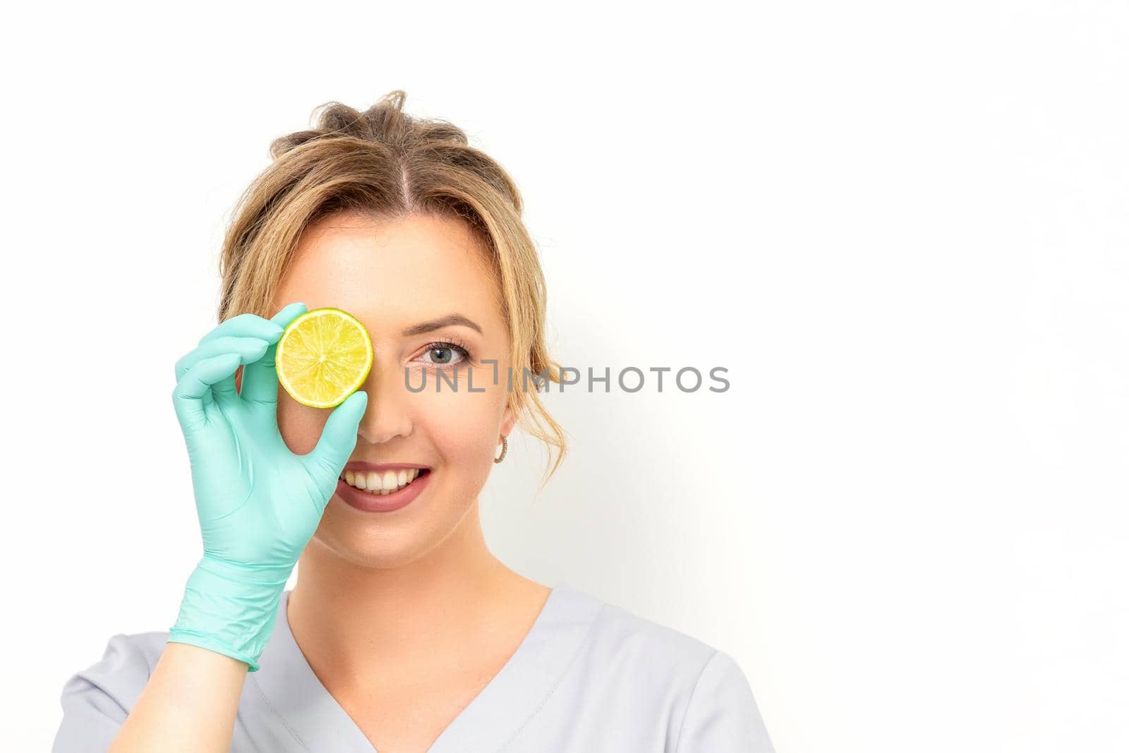 Portrait of young caucasian smiling female beautician covering eye with a lime slice wearing gloves over a white background