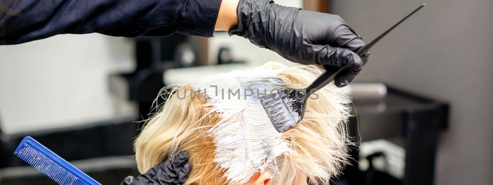 Coloring white hair with hair dye and brush by hands of hairstylist for the young caucasian blonde woman at a hair salon, close up