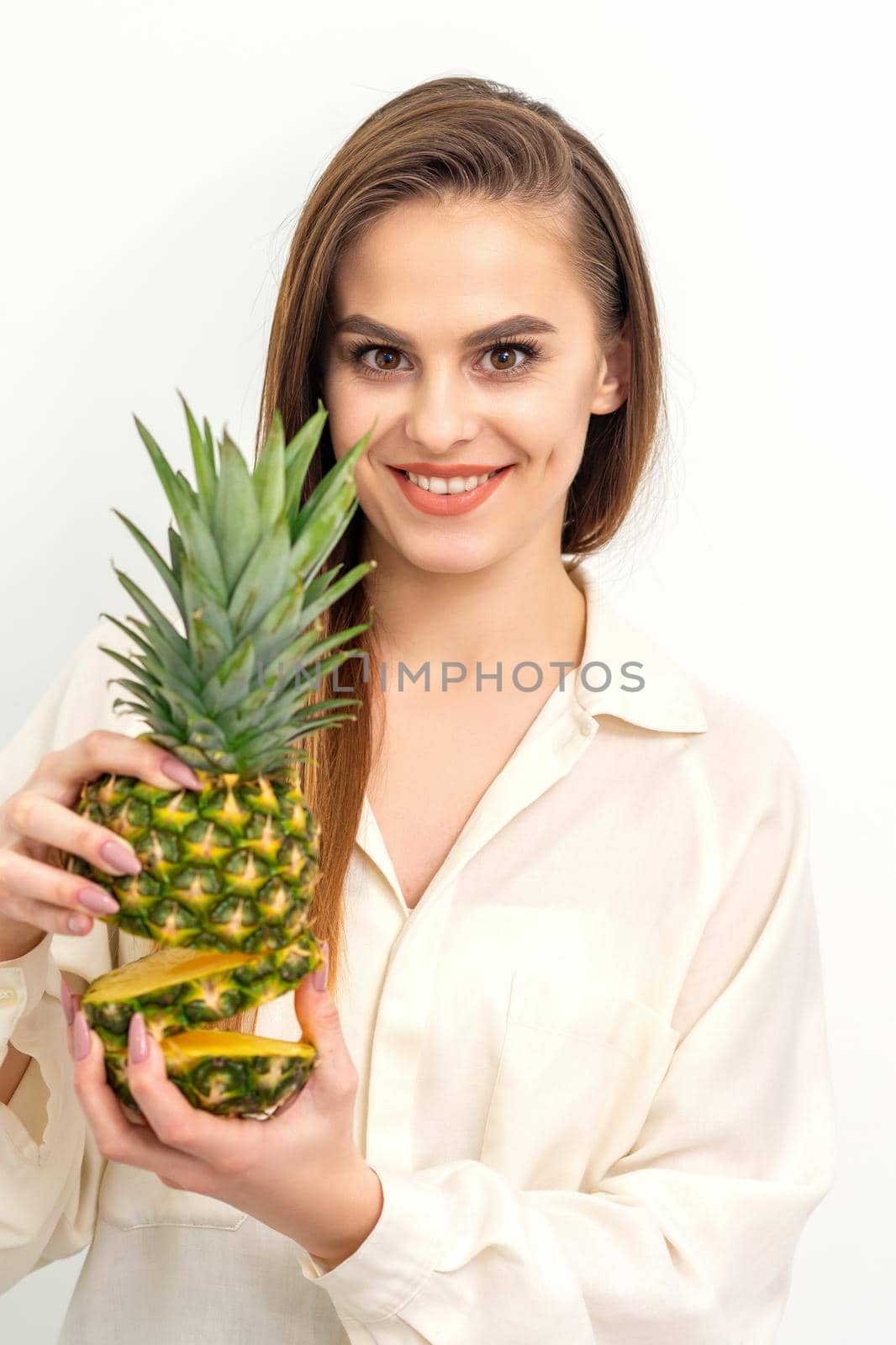 Beautiful young Caucasian woman holding pineapple and smiling, wearing a white shirt over white background