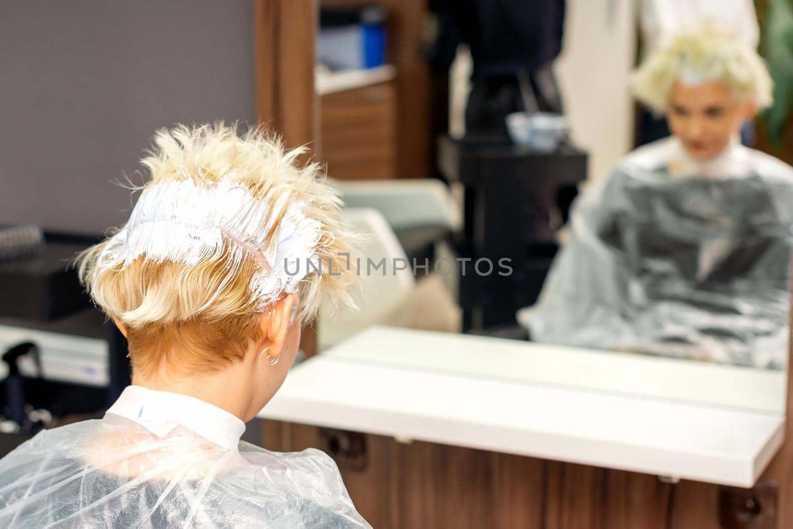 Coloring white hair with hair dye of the young caucasian blonde woman sitting at a hair salon, close up