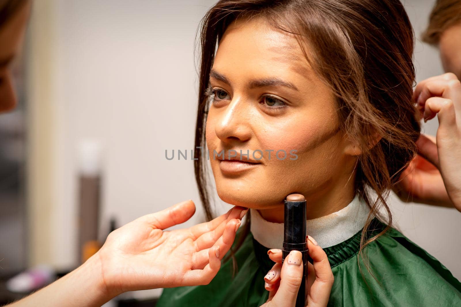 Beautiful young brunette woman receiving makeup with stick concealer on her face in a beauty salon