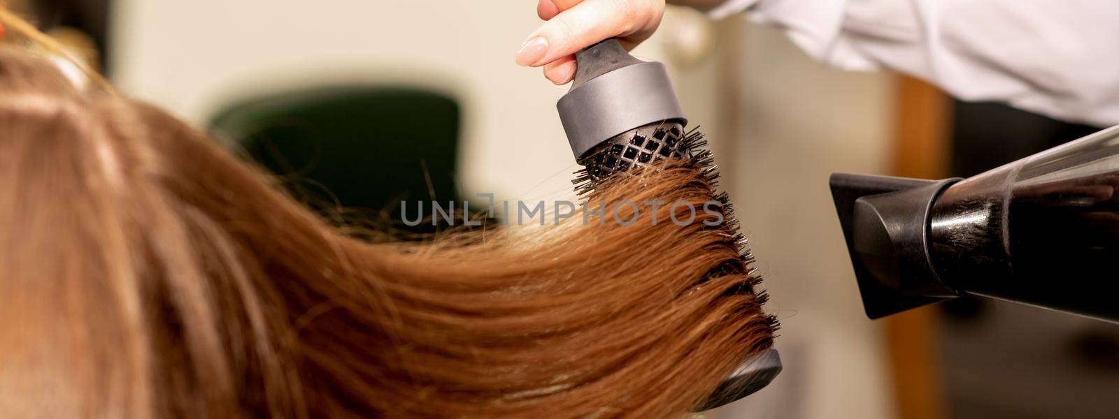 A hairdresser is drying long brown hair with a hairdryer and round brush in a beauty salon. by okskukuruza