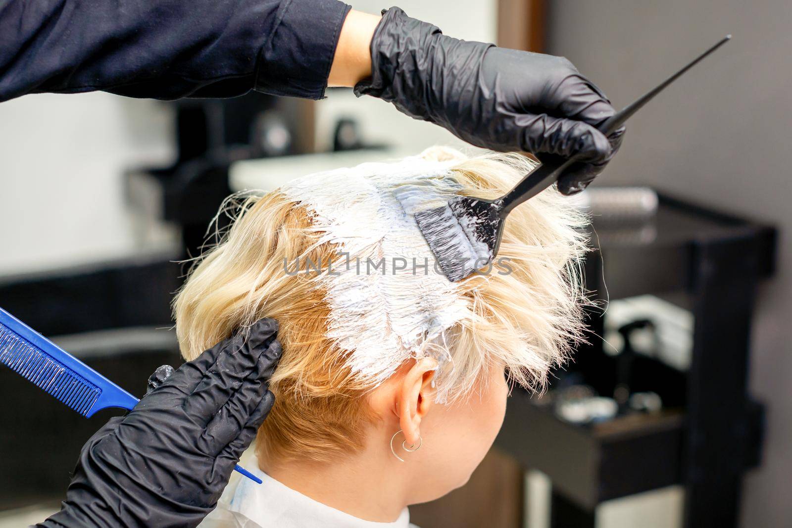 Coloring white hair with hair dye and brush by hands of hairstylist for the young caucasian blonde woman at a hair salon, close up